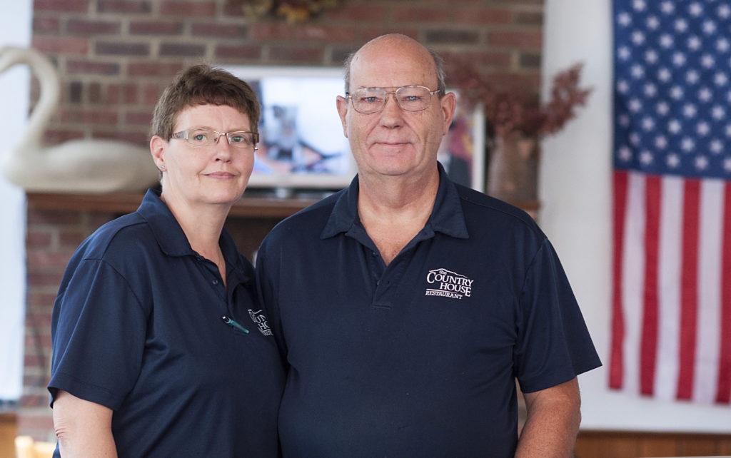 Don and Donna Pelton wear blue shirts and pose by the American Flag at The Country House restaurant