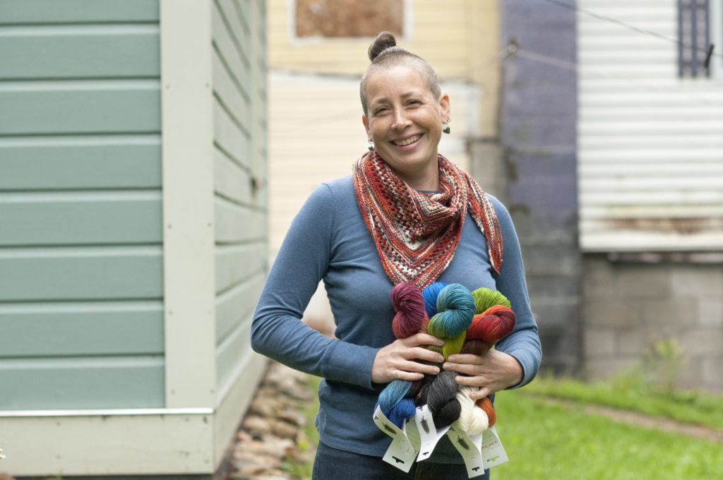 Monica MacNeille of Round Mountain fibers wears a blue sweater and jeans and poses with her hand-dyed, hand-spun yarns, holding skeins in a variety of blue, green and red shades