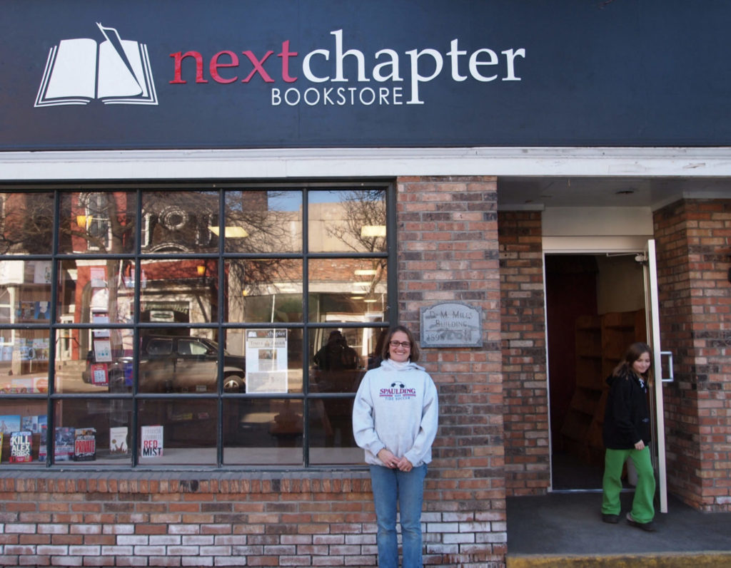Cynthia Duprey of Next Chapter bookstore in Barre stands in front of her building, smiling, wearing a gray sweatshirt and jeans