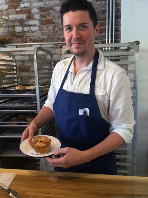 Employee of Piecemeal Pies poses with one of his pies, wearing a blue apron and white chef's coat while standing in his kitchen